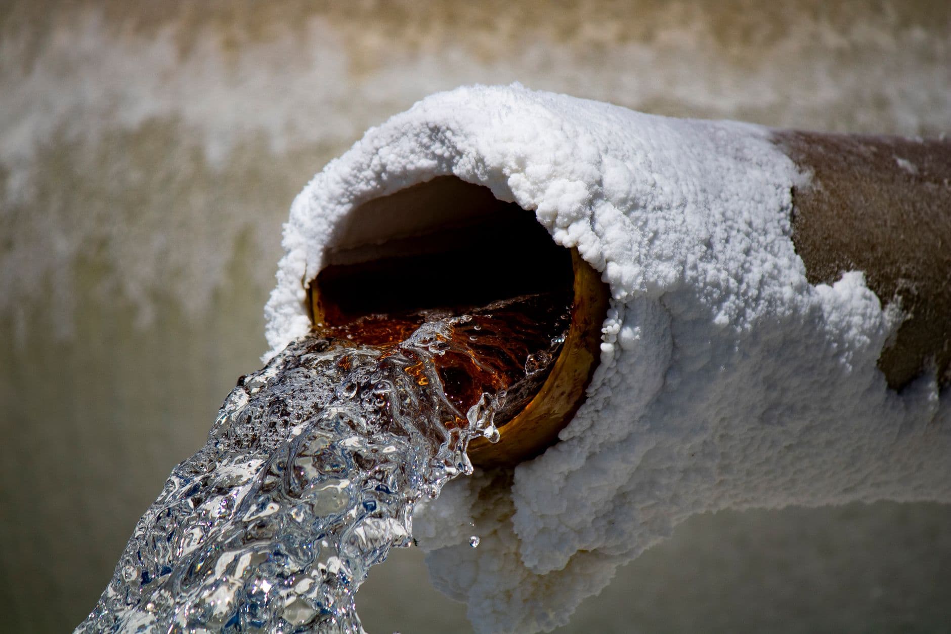 Water running from a pipe whose mouth is thickly encrusted with white mineral scale, the bore inside stained orange.