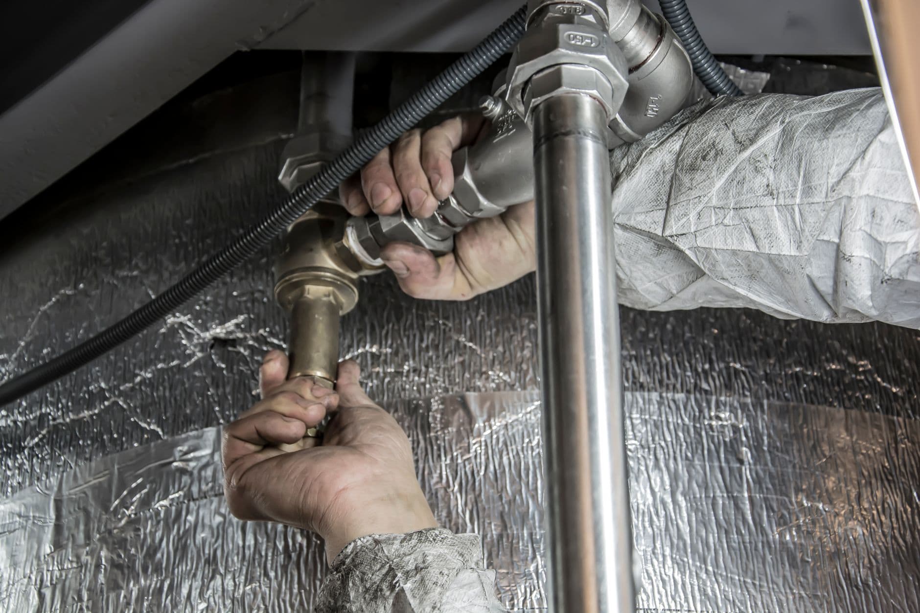 Two hands tightening a threaded union on brass and steel pipework beside insulated ducting.