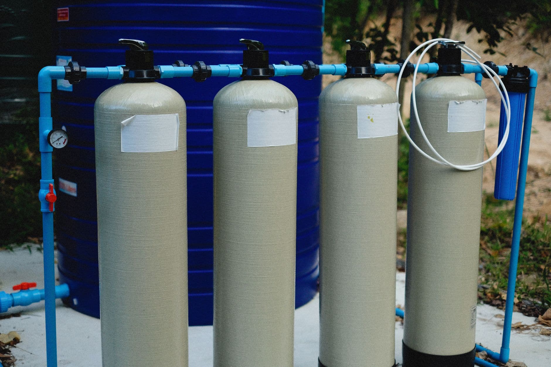 Four water filtration vessels with valves and a pressure gauge, plumbed in blue pipework in front of a large water storage tank.